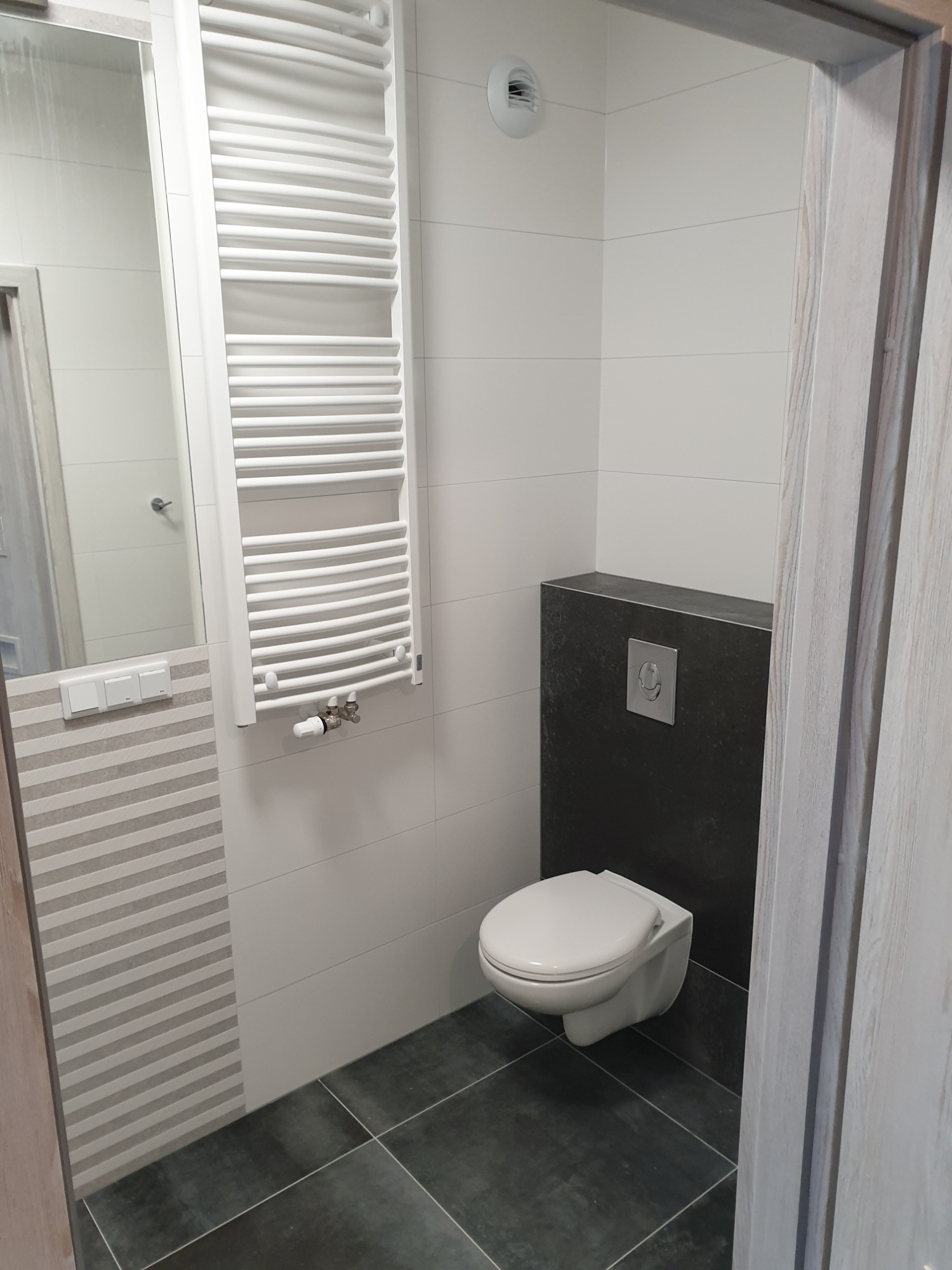 Nowoczesna łazienka z wiszącą toaletą na tle ciemnoszarej ściany, białe płytki, grzejnik drabinkowy i lustro z szarymi pasami. Ciemna podłoga.