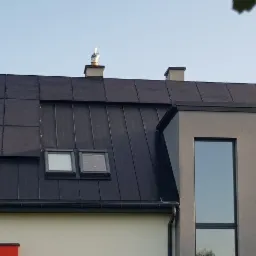 Czarny dach z blachy na nowoczesnym domu z panelami fotowoltaicznymi w górnej części połaci, widoczne okna dachowe i kominy.