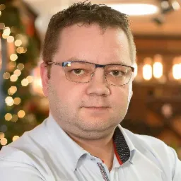 Portret mężczyzny w okularach, z delikatnym uśmiechem, na rozmytym tle z dekoracyjnymi światełkami. Ubrany w jasną koszulę.