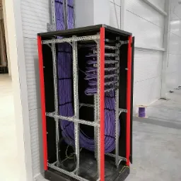 Szafa rack z uporządkowanymi fioletowymi kablami sieciowymi, widoczny fragment kabli prowadzonych w korytkach na ścianie, w tle szpula z zapasem kabla.