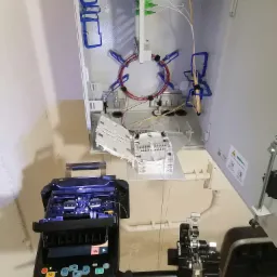 Otwarta szafka telekomunikacyjna z widocznymi kablami światłowodowymi, złączami i narzędziami do spawania światłowodów, w trakcie prac instalacyjnych.