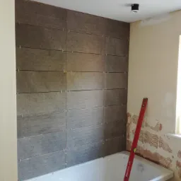 Montaż szarych płytek imitujących drewno na ścianie nad wanną w łazience w Tarnobrzegu, widoczny poziomica oparta o ścianę i krzyżyki dystansowe między płytkami.