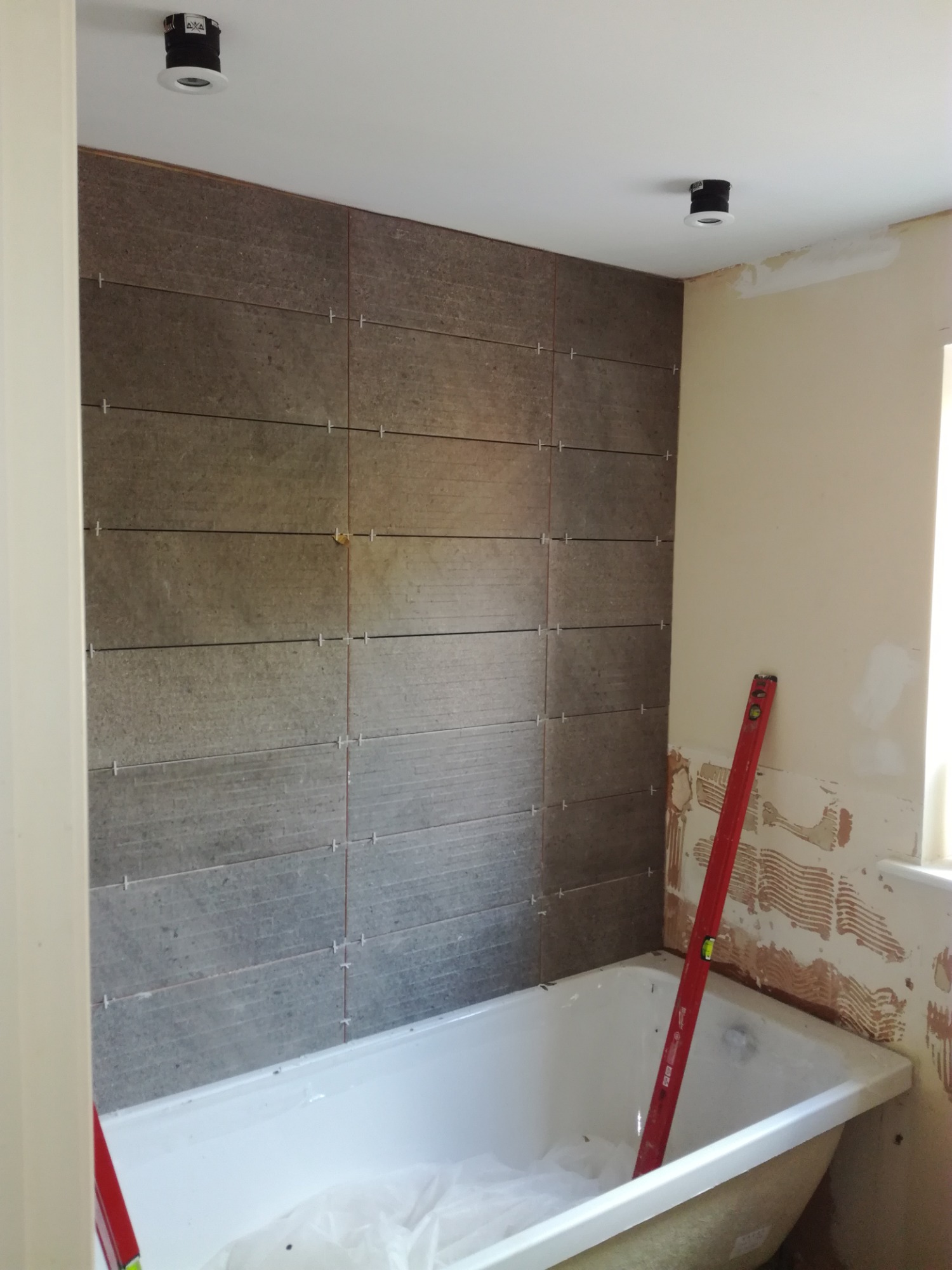 Montaż szarych płytek imitujących drewno na ścianie nad wanną w łazience w Tarnobrzegu, widoczny poziomica oparta o ścianę i krzyżyki dystansowe między płytkami.