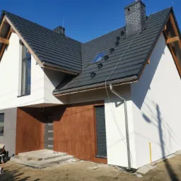 Nowoczesny dom jednorodzinny z białymi ścianami i brązowym akcentem przy wejściu, widoczna nowa dachówka i orynnowanie, teren wokół domu jeszcze niezagospodarowany, w tle błękitne niebo.