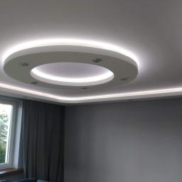 Zmaźlok - Nowoczesny sufit podwieszany z okrągłym elementem centralnym i oświetleniem LED wzdłuż krawędzi, widok z dołu na szarą ścianę i okno z zasłoną.