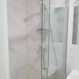 Zmaźlok - Nowoczesna kabina prysznicowa z przesuwnymi drzwiami, wykończona białymi płytkami imitującymi marmur, z widoczną armaturą i półką na kosmetyki.