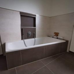 Zmaźlok - Biała wanna obudowana brązowymi płytkami w łazience z beżowymi ścianami, widoczna bateria i wnęka w ścianie. Na wannie stoi kartonowe pudełko i drewniana figurka misia.