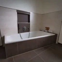 Biała wanna obudowana brązowymi płytkami w łazience z beżowymi ścianami, widoczna bateria i wnęka w ścianie. Na wannie stoi kartonowe pudełko i drewniana figurka misia.