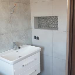 Zmaźlok - Nowoczesna łazienka w odcieniach szarości z białą szafką pod umywalką z dwoma szufladami, ozdobną wnęką wyłożoną mozaiką i wystającymi przewodami elektrycznymi na ścianie.