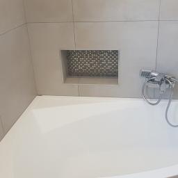 Zmaźlok - Biała wanna narożna z chromowaną baterią i słuchawką prysznicową na tle szarych płytek ceramicznych, z wnęką wyłożoną mozaiką w kształcie drobnej kratki.