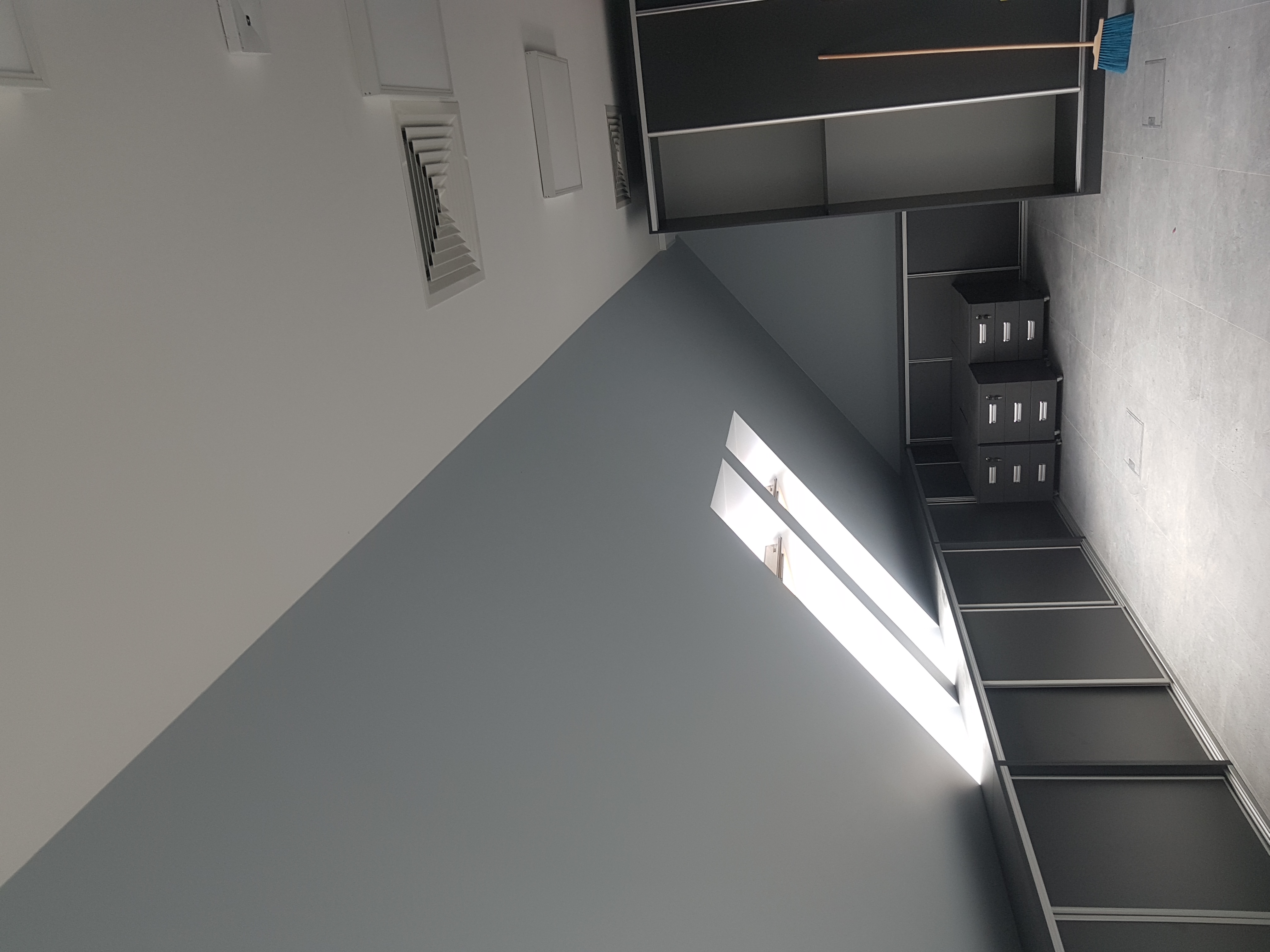 Wnętrze biura z szarą ścianą, oknem z jasnym światłem, czarnymi szafami z przesuwnymi drzwiami i niebieską miotłą opartą o szafę.