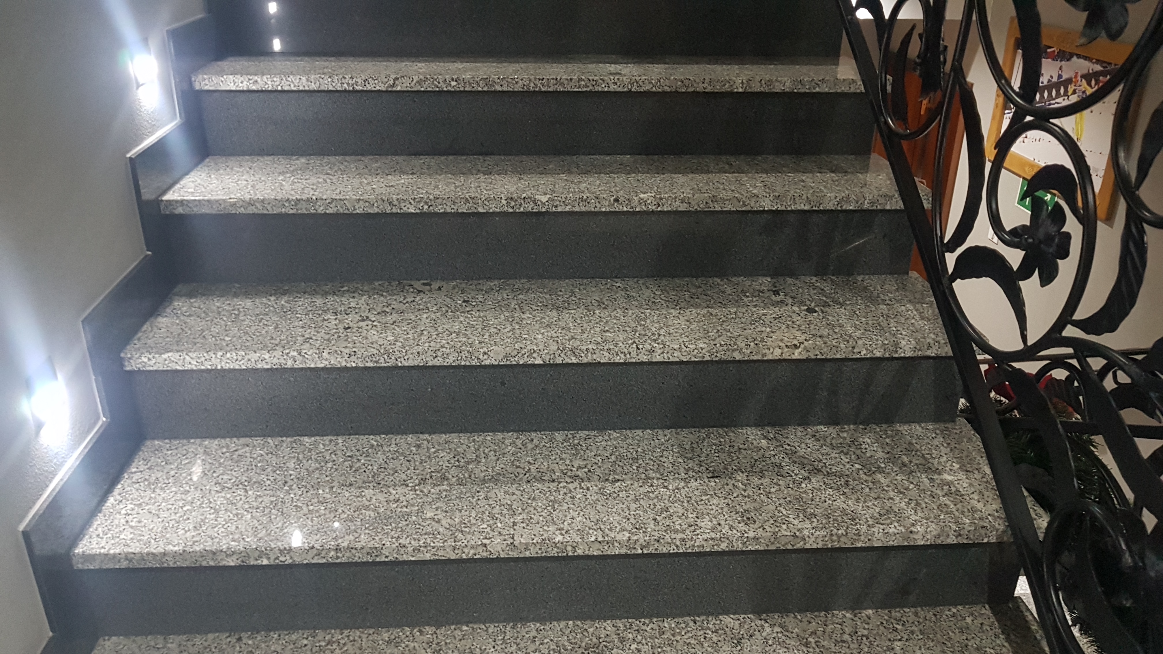Granitowe schody z czarnymi podstopnicami, oświetlone punktowo, z dekoracyjną, kutą balustradą.