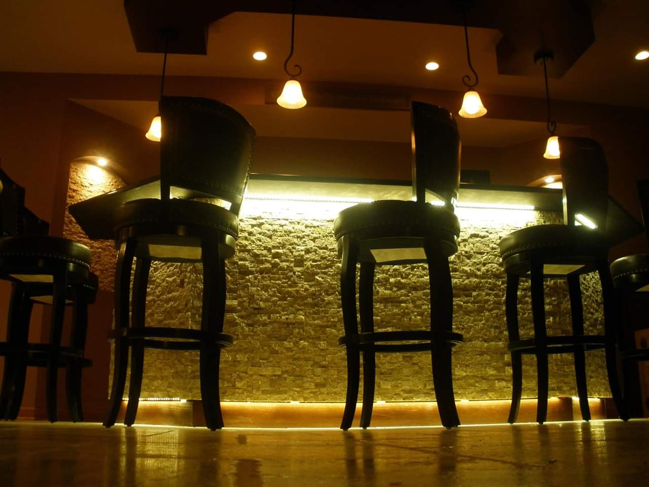 Oświetlony bar z kamienną ścianą i wysokimi, skórzanymi krzesłami barowymi, widok z niskiej perspektywy podkreśla refleksy na polerowanej podłodze.
