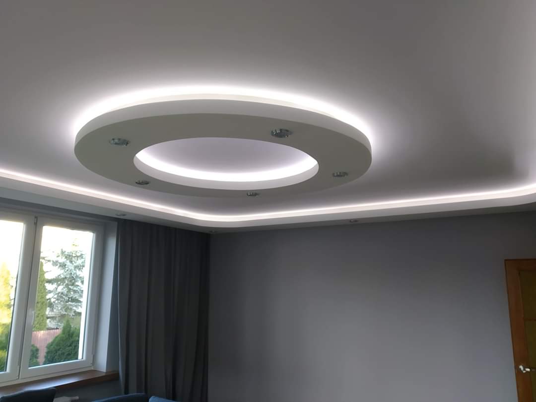 Nowoczesny sufit podwieszany z okrągłym elementem centralnym i oświetleniem LED wzdłuż krawędzi, widok z dołu na szarą ścianę i okno z zasłoną.