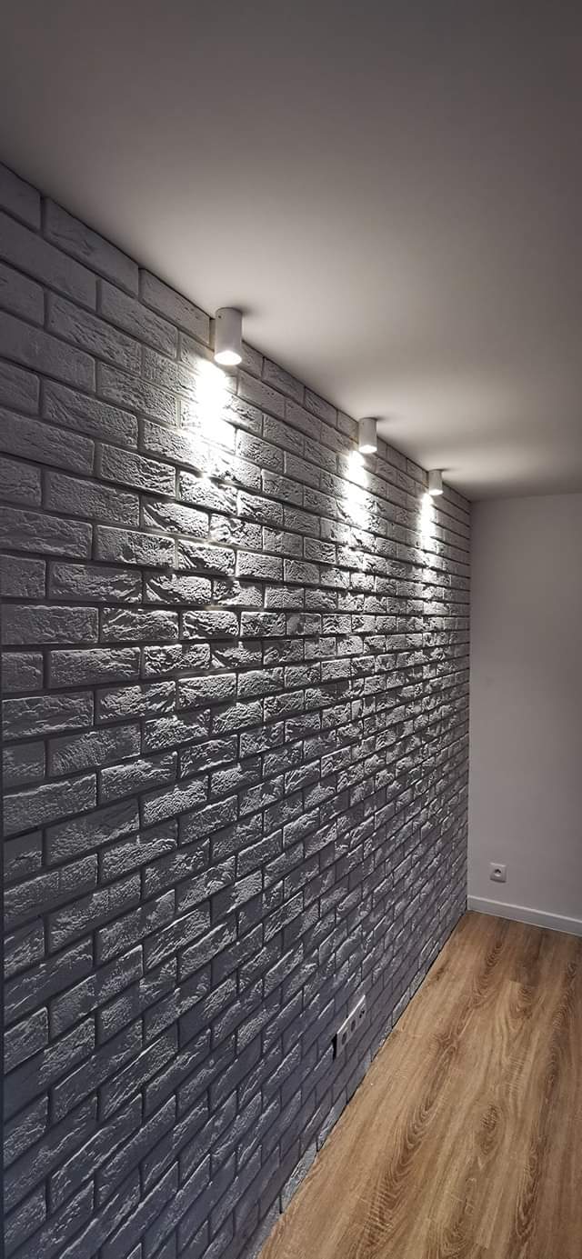 Ściana wykończona szarą cegłą dekoracyjną, oświetlona trzema punktowymi lampami sufitowymi, z widoczną drewnopodobną podłogą i białym narożnikiem ściany.