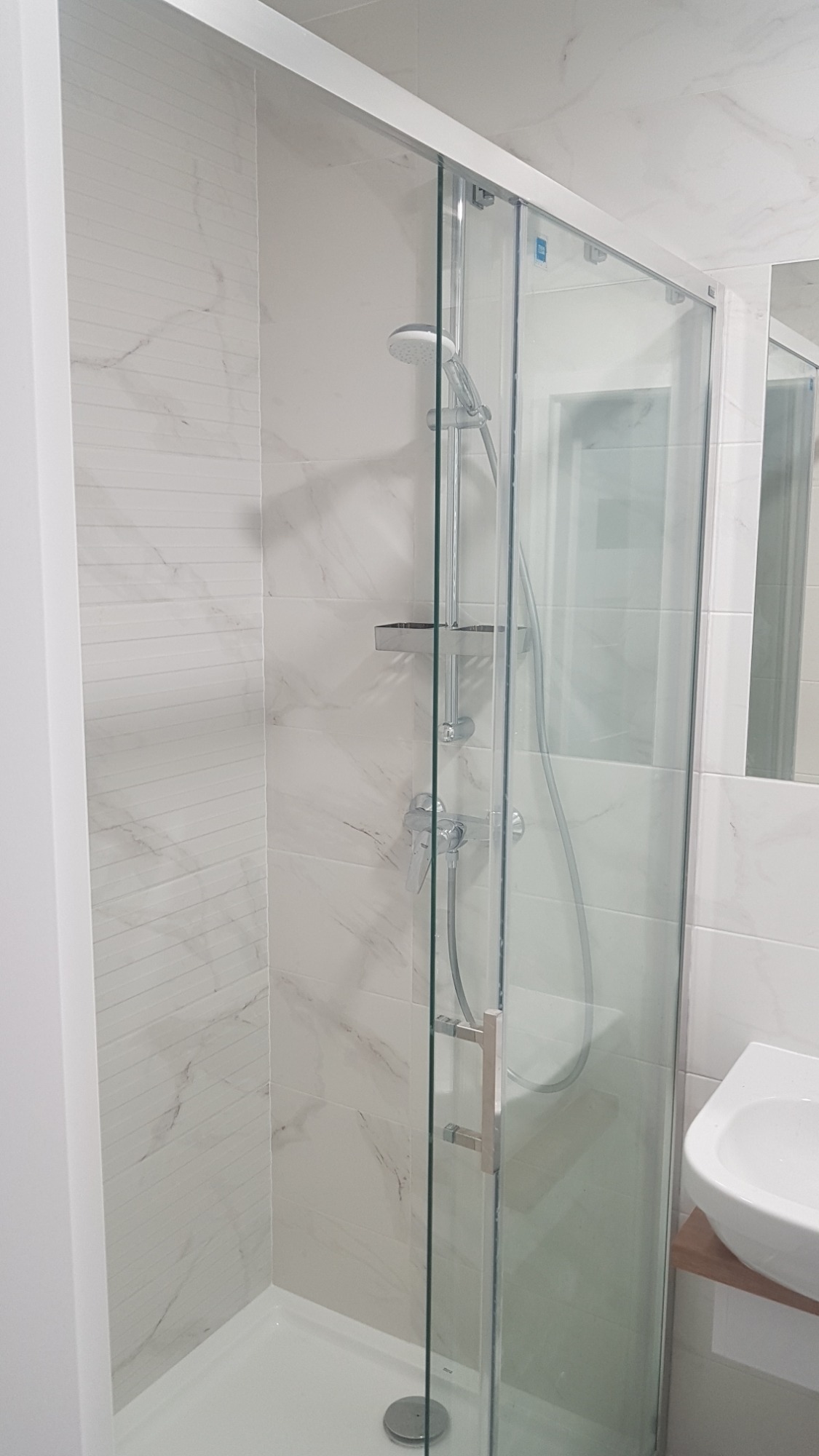 Nowoczesna kabina prysznicowa z przesuwnymi drzwiami, wykończona białymi płytkami imitującymi marmur, z widoczną armaturą i półką na kosmetyki.