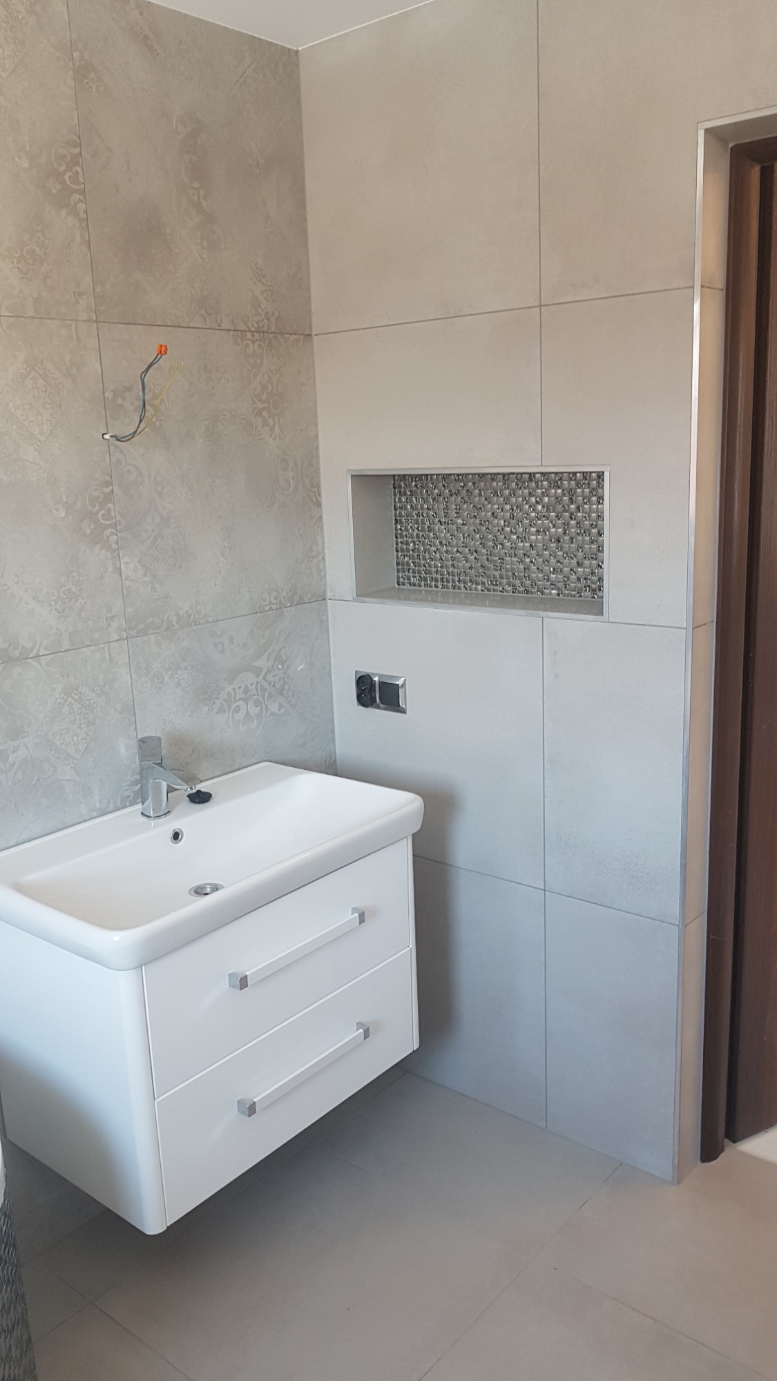 Nowoczesna łazienka w odcieniach szarości z białą szafką pod umywalką z dwoma szufladami, ozdobną wnęką wyłożoną mozaiką i wystającymi przewodami elektrycznymi na ścianie.