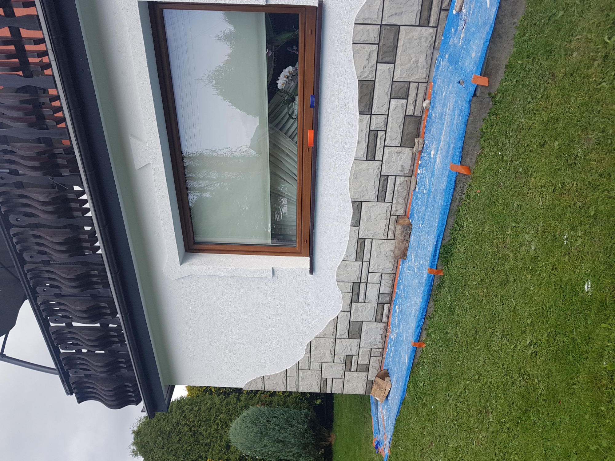 Fragment elewacji domu z oknem, częściowo wykończony kamieniem dekoracyjnym, zabezpieczony folią malarską na trawniku.