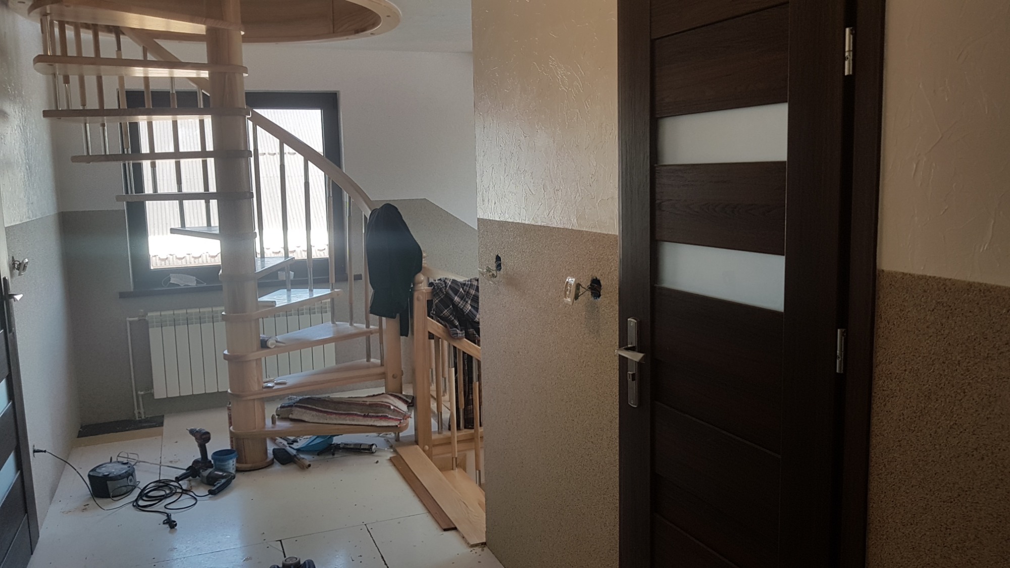 Wnętrze w trakcie prac wykończeniowych: drewniane schody spiralne, częściowo wykończona ściana z widocznymi puszkami elektrycznymi, drzwi z ciemnego drewna z przeszkleniami, narzędzia na podłodze.