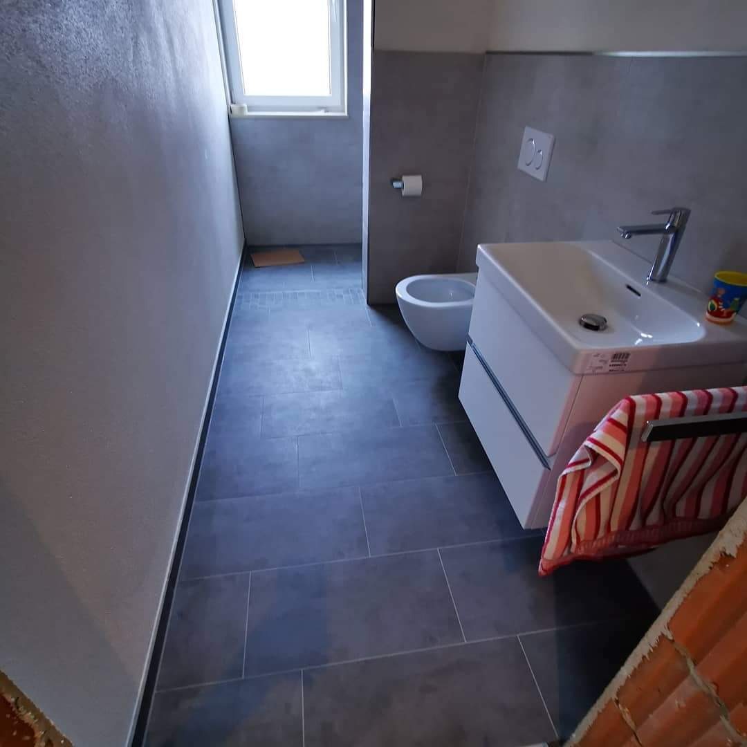 Nowoczesna łazienka z szarymi płytkami podłogowymi i ściennymi, białą umywalką z szafką i bidetem, widok z perspektywy wejścia, częściowo widoczna ściana z cegieł.