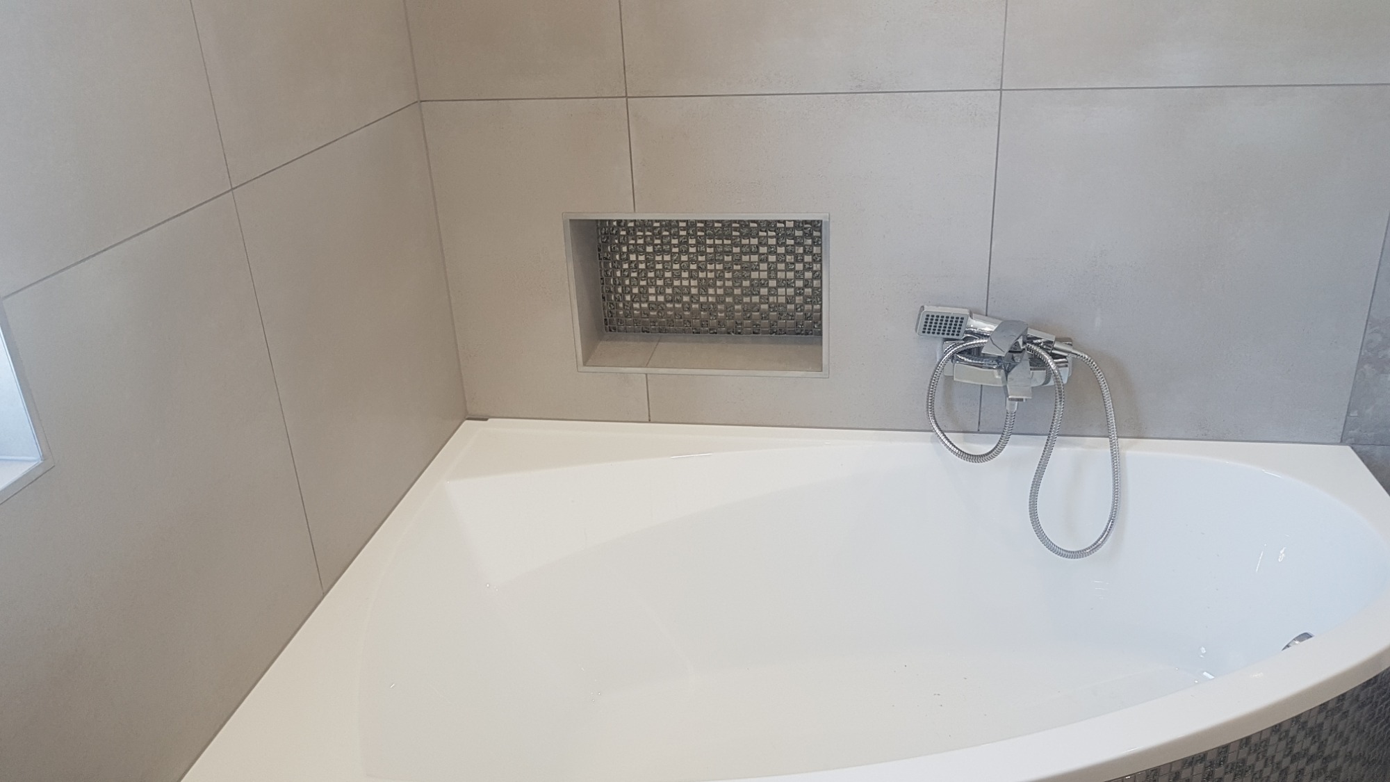 Biała wanna narożna z chromowaną baterią i słuchawką prysznicową na tle szarych płytek ceramicznych, z wnęką wyłożoną mozaiką w kształcie drobnej kratki.