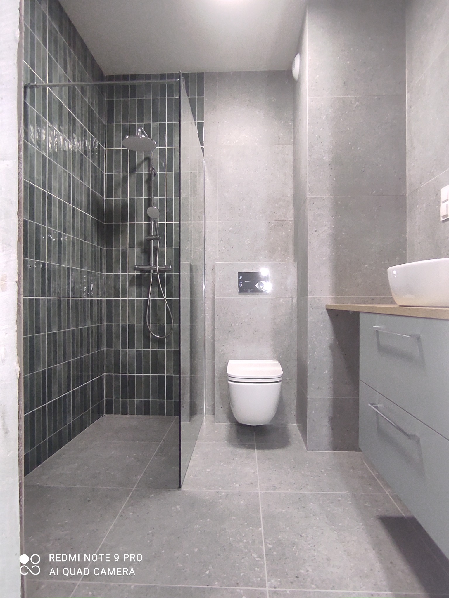 Nowoczesna łazienka z kabiną typu walk-in, wykończona szarymi płytkami i zieloną mozaiką w strefie prysznica. Uwagę zwraca minimalistyczna armatura i podwieszana toaleta.