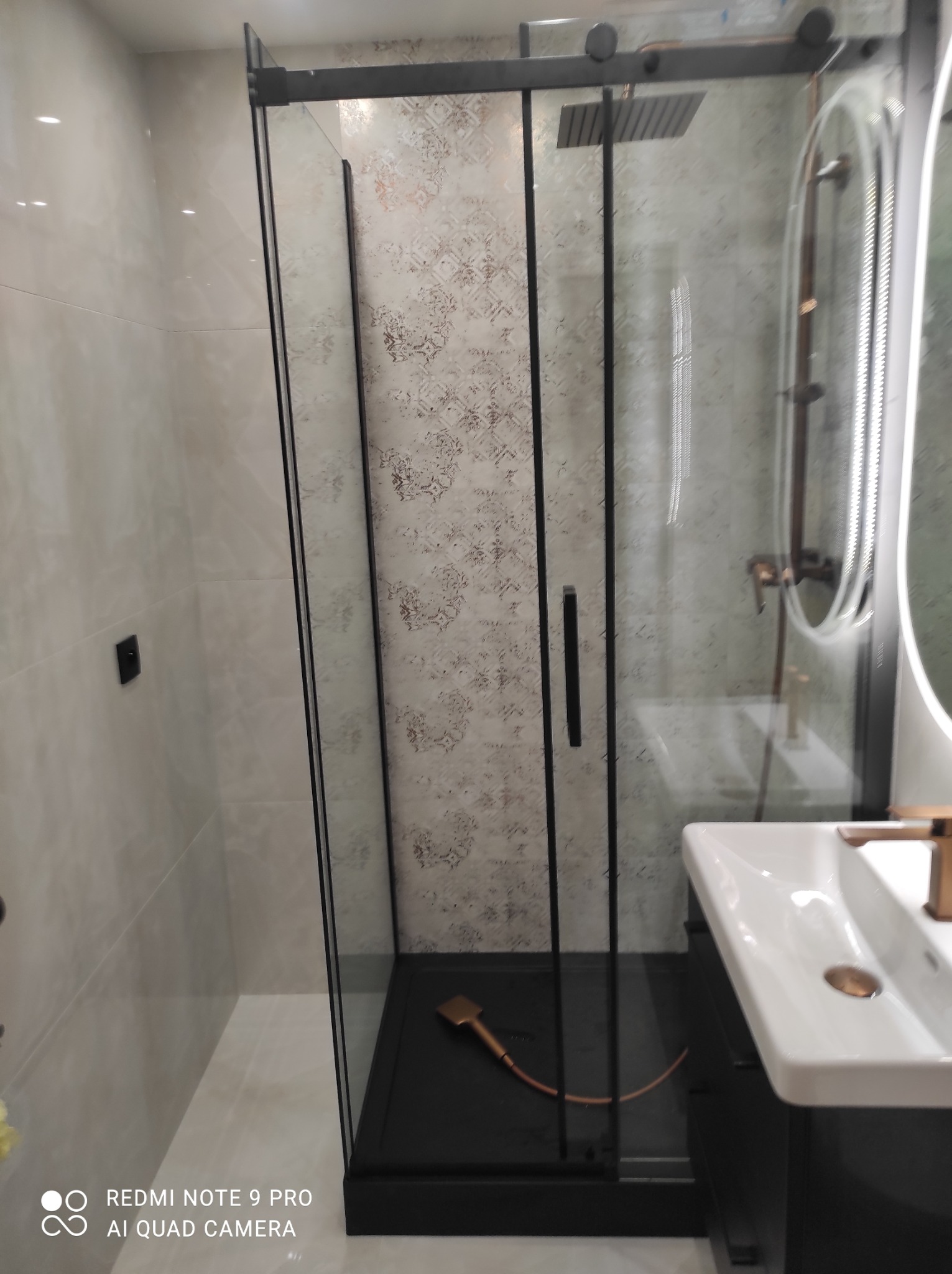 Nowoczesna kabina prysznicowa z czarnymi profilami i deszczownicą, umywalka z baterią w kolorze miedzi, płytki z delikatnym wzorem. Wykończona łazienka.