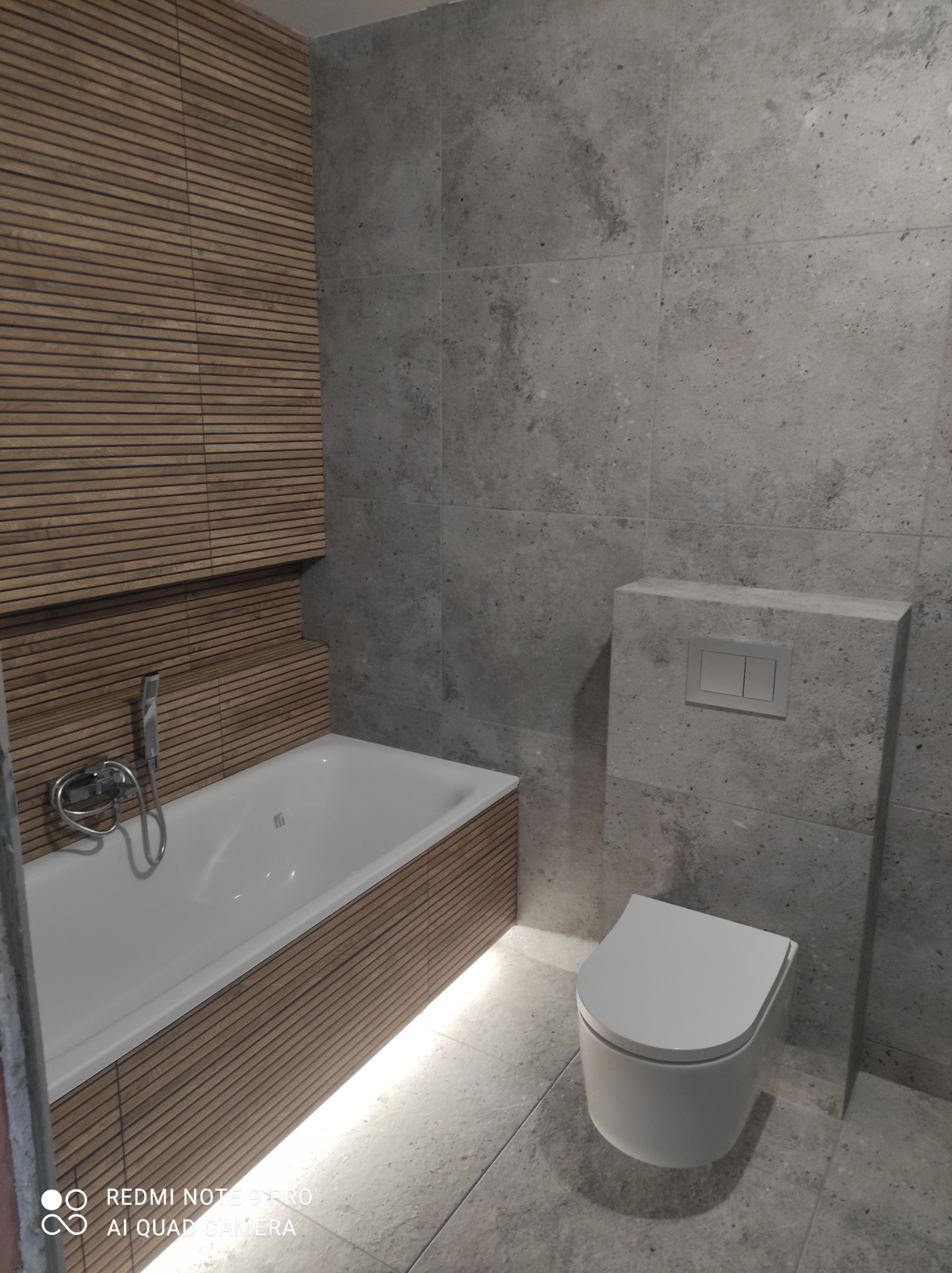 Nowoczesna łazienka z wanną obudowaną drewnianymi panelami i podświetleniem LED, oraz toaletą w zabudowie z szarych płytek imitujących kamień.