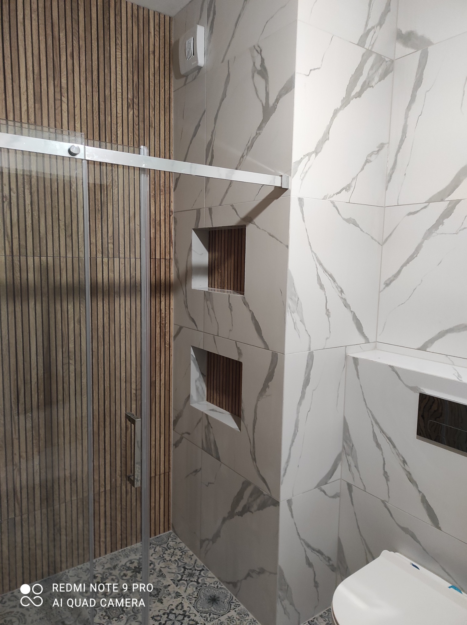 Nowoczesna łazienka po remoncie: kabina prysznicowa ze szklanymi drzwiami, marmurowe płytki z szarymi żyłami, drewniane panele ścienne i dekoracyjna podłoga.