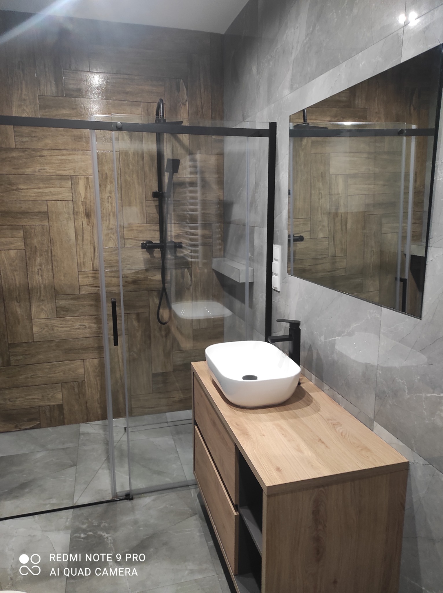 Nowoczesna łazienka z kabiną prysznicową typu walk-in, drewnianą szafką pod umywalką i szarymi płytkami imitującymi marmur na ścianach. Czarna armatura.