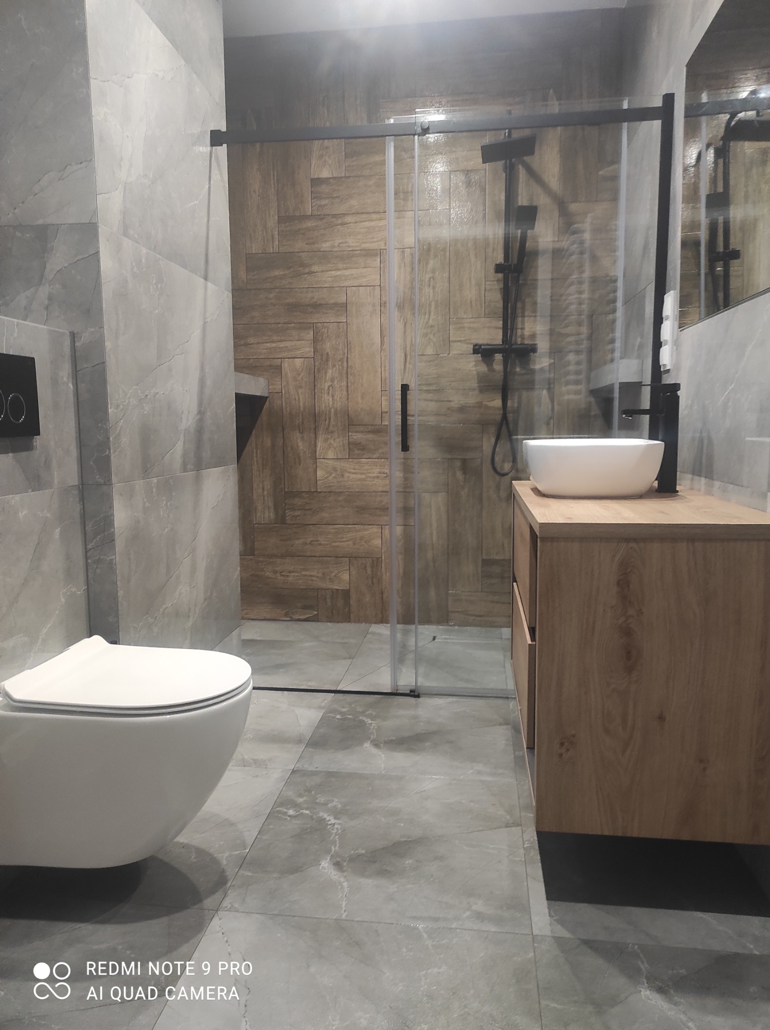 Nowoczesna łazienka po remoncie: szare płytki imitujące marmur, drewnopodobne panele na ścianie prysznica, wisząca toaleta i szafka z umywalką.