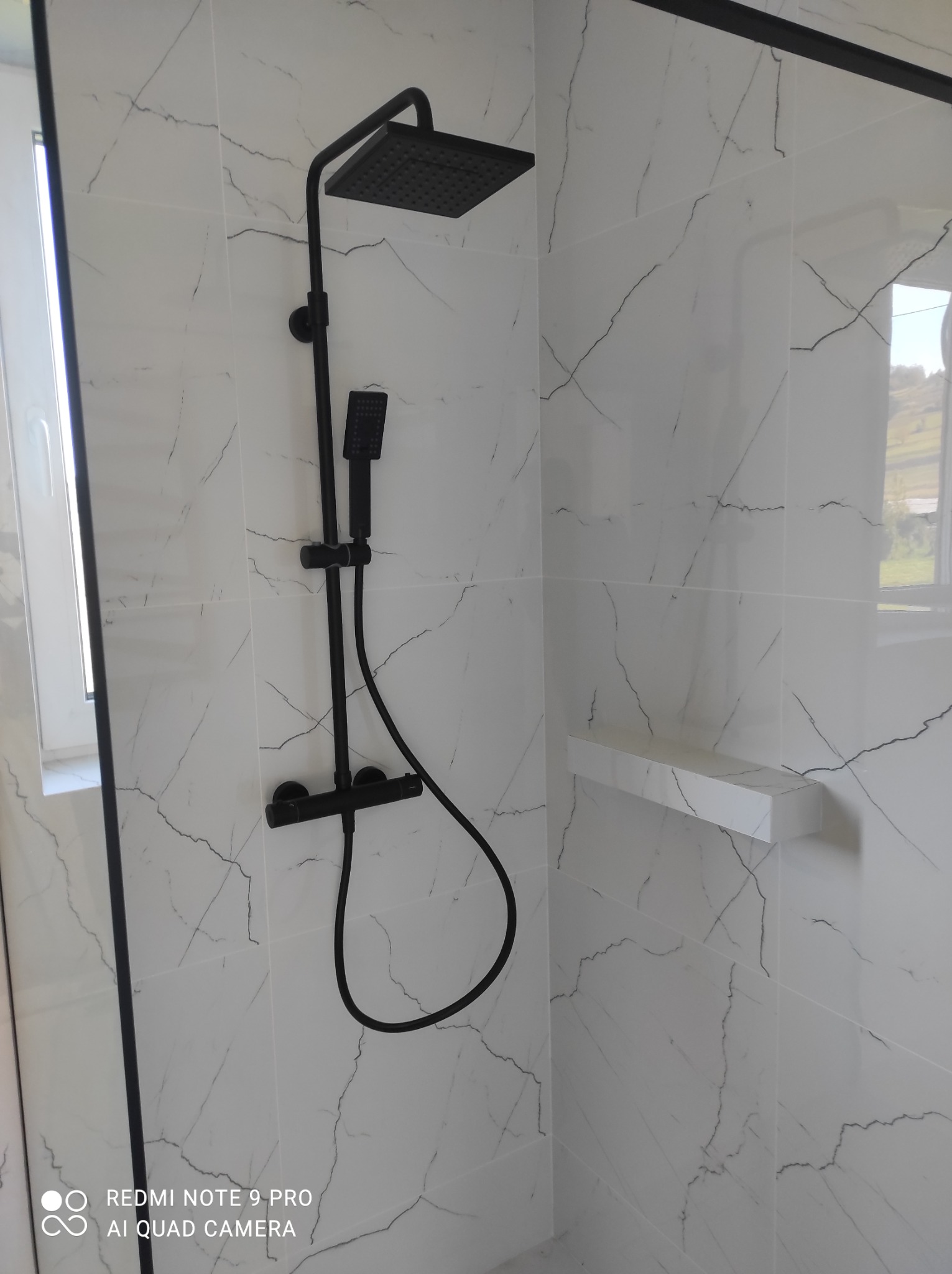 Nowoczesna kabina prysznicowa z czarną armaturą na tle białych płytek imitujących marmur. Minimalistyczny design, widoczna półka wewnątrz kabiny.