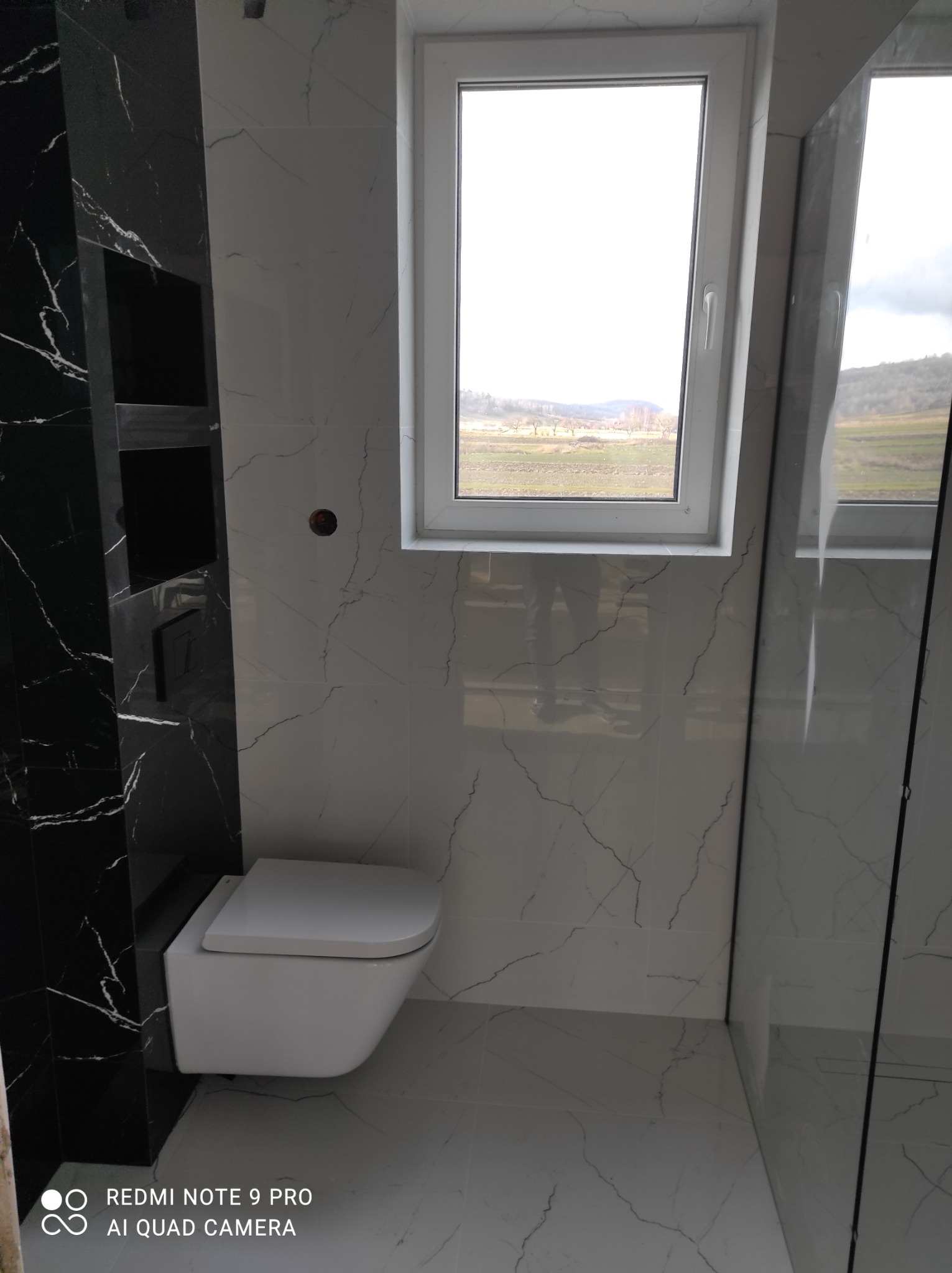 Nowoczesna łazienka z czarno-białymi płytkami imitującymi marmur, podwieszana toaleta i okno z widokiem na krajobraz. Minimalistyczny design.