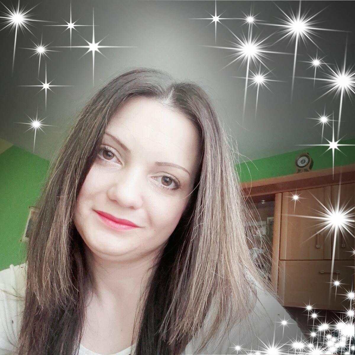 Portret uśmiechniętej kobiety z długimi brązowymi włosami i czerwonymi ustami, z efektem rozbłysku gwiazd na obrzeżach zdjęcia, w tle zielona ściana i drewniana szafka z zegarem.