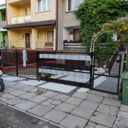 Nowa, czarna brama panelowa z jasnoszarymi wypełnieniami poziomymi, zamontowana przed domem z kostki brukowej, widoczny fragment motocykla po lewej stronie i skrzynka na listy po prawej.