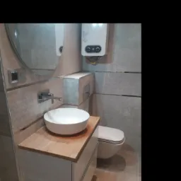 Nowoczesna łazienka z owalną umywalką nablatową na szafce z drewnianym blatem, szarą płytką na ścianach z metalowymi listwami dekoracyjnymi, wiszącą toaletą i podgrzewaczem wody.