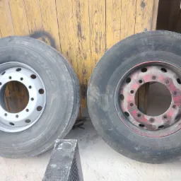 Dwa koła ciężarowe: jedno z felgą pomalowaną na srebrno, drugie z felgą w kolorze czerwonym, stojące na betonowej powierzchni przed drewnianą ścianą.