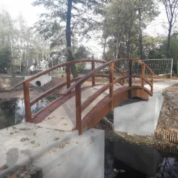 Drewniany mostek łukowy nad strumieniem w parku, świeżo pomalowany, widoczne zabezpieczenia budowlane w tle, jesienne liście na betonowej podstawie.