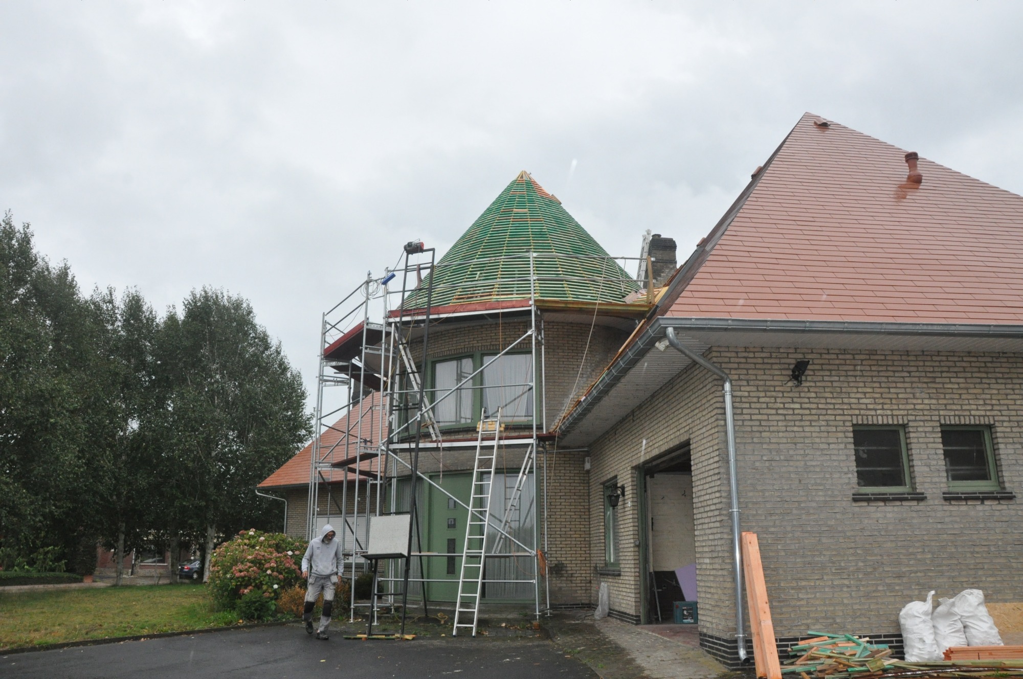 Budowa dachu z zielonymi elementami na okrągłej wieży domu, widoczne rusztowania i pracownik w oddali.