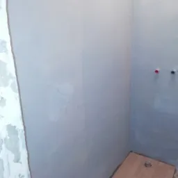 Świeżo wyszpachlowana ściana w rogu pomieszczenia, z widocznymi śladami po szpachlowaniu oraz fragmentem podłogi z ułożonymi panelami w odcieniach brązu, przygotowana do malowania.