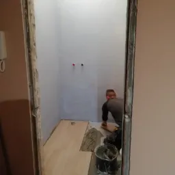 Układanie paneli podłogowych w małym pomieszczeniu, widok przez futrynę bez drzwi, pracownik docina panel, obok wiadro z klejem i narzędzia.