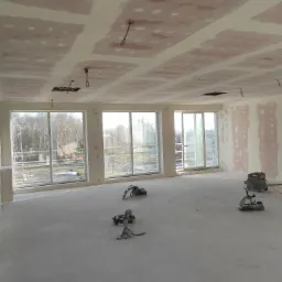 Wnętrze w trakcie prac wykończeniowych, widoczne płyty kartonowo-gipsowe na suficie i ścianach z widocznymi łączeniami szpachlowanymi, na podłodze porozstawiane narzędzia i odkurzacz przemysłowy...