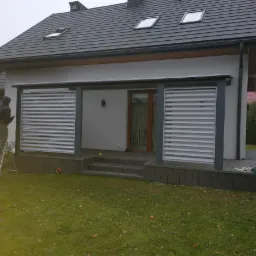 Nowoczesna konstrukcja z poziomych, białych paneli na tarasie z kompozytowych desek, pracownik na drabinie w trakcie montażu, widoczne narzędzia i skrzynka narzędziowa, dom z szarym dachem w tle.