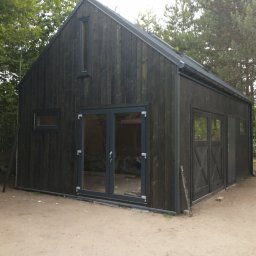 Robkalwik - Nowoczesny, ciemny budynek z drewnianą elewacją i dwuspadowym dachem, z widocznymi oknami i drzwiami z szarymi ramami, otoczony drzewami i piaszczystym terenem.