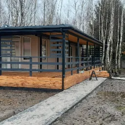 Nowoczesny, parterowy dom z drewnianym tarasem i szarą balustradą, otoczony brzozowym lasem i błotnistym terenem.