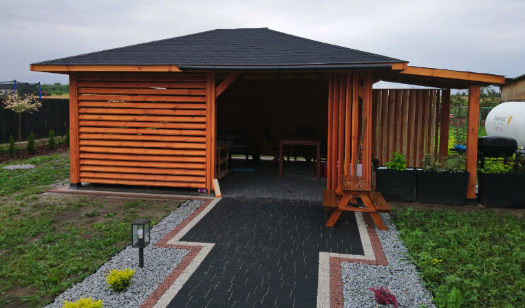 Drewniana konstrukcja z czarnym dachem, częściowo osłonięta poziomymi panelami, z widocznym wnętrzem i stołem, otoczona kostką brukową i zielenią, z lampą ogrodową i zbiornikiem na gaz w tle.