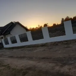 Nowoczesne ogrodzenie panelowe, szare poziome lamele między białymi słupkami, na tle domu i zachodzącego słońca.