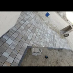 Ułożona szara kostka brukowa na podjeździe, widok z góry na fragment posesji w trakcie prac wykończeniowych, widoczne narzędzia i materiały budowlane.