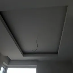 Wykończony sufit z obniżonym kwadratowym panelem, widoczne wystające kable oświetleniowe, surowe ściany i okno w tle.