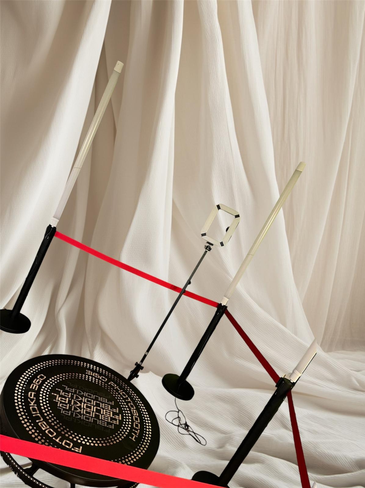 Elementy składowe fotobudki 360 stopni z czerwonymi taśmami na tle drapowanej białej tkaniny.